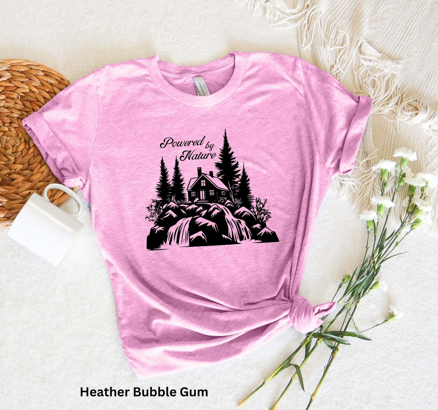 Light Pink T-shirt with the phrase 'Powered by Nature,' featuring a detailed cabin in the woods, a cascading waterfall, and trees—perfect for outdoor and adventure lovers