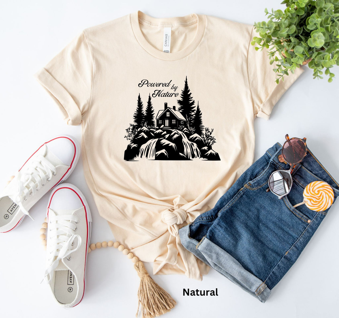 T-shirt in natural color with the phrase 'Powered by Nature,' featuring a detailed cabin in the woods, a cascading waterfall, and trees—perfect for outdoor and adventure lovers
