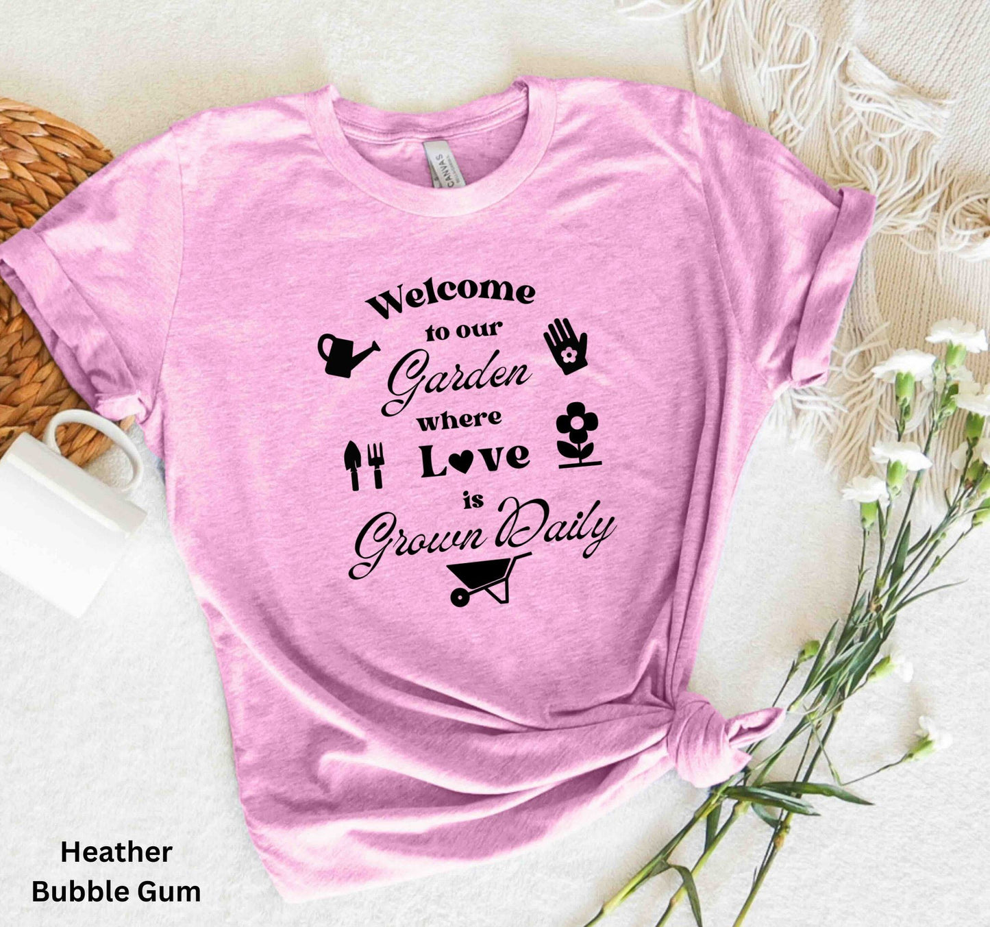 Light Pink T-shirt with the phrase 'Welcome to our Garden where love is grown daily,' featuring gardening tools, a heart, a wheelbarrow, a flower, gloves, and a watering jug.
