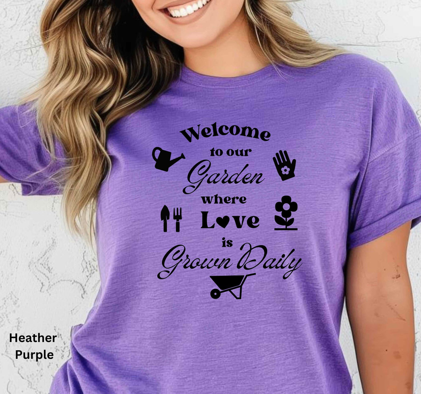 Purple T-shirt with the phrase 'Welcome to our Garden where love is grown daily,' featuring gardening tools, a heart, a wheelbarrow, a flower, gloves, and a watering jug.