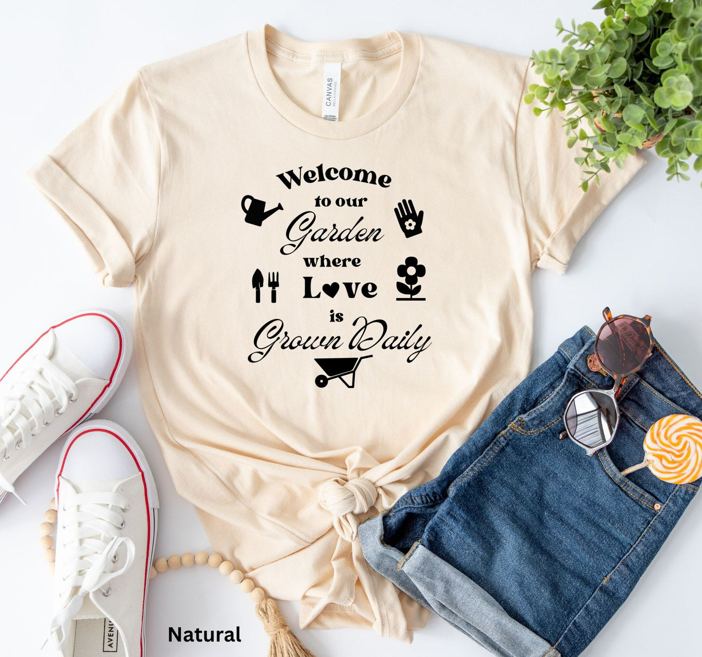 T-shirt in natural color with the phrase 'Welcome to our Garden where love is grown daily,' featuring gardening tools, a heart, a wheelbarrow, a flower, gloves, and a watering jug.