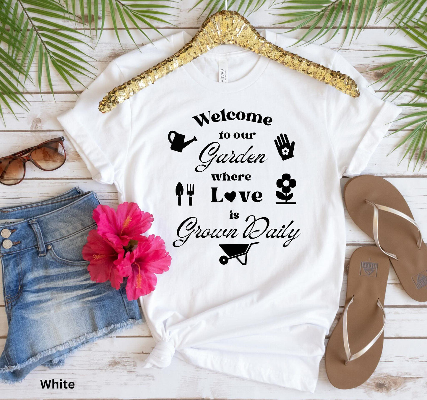 White T-shirt with the phrase 'Welcome to our Garden where love is grown daily,' featuring gardening tools, a heart, a wheelbarrow, a flower, gloves, and a watering jug.